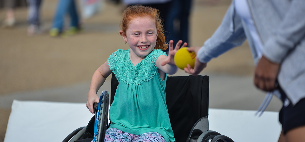 A girl in a wheelchair takes a tennis ball from a coach