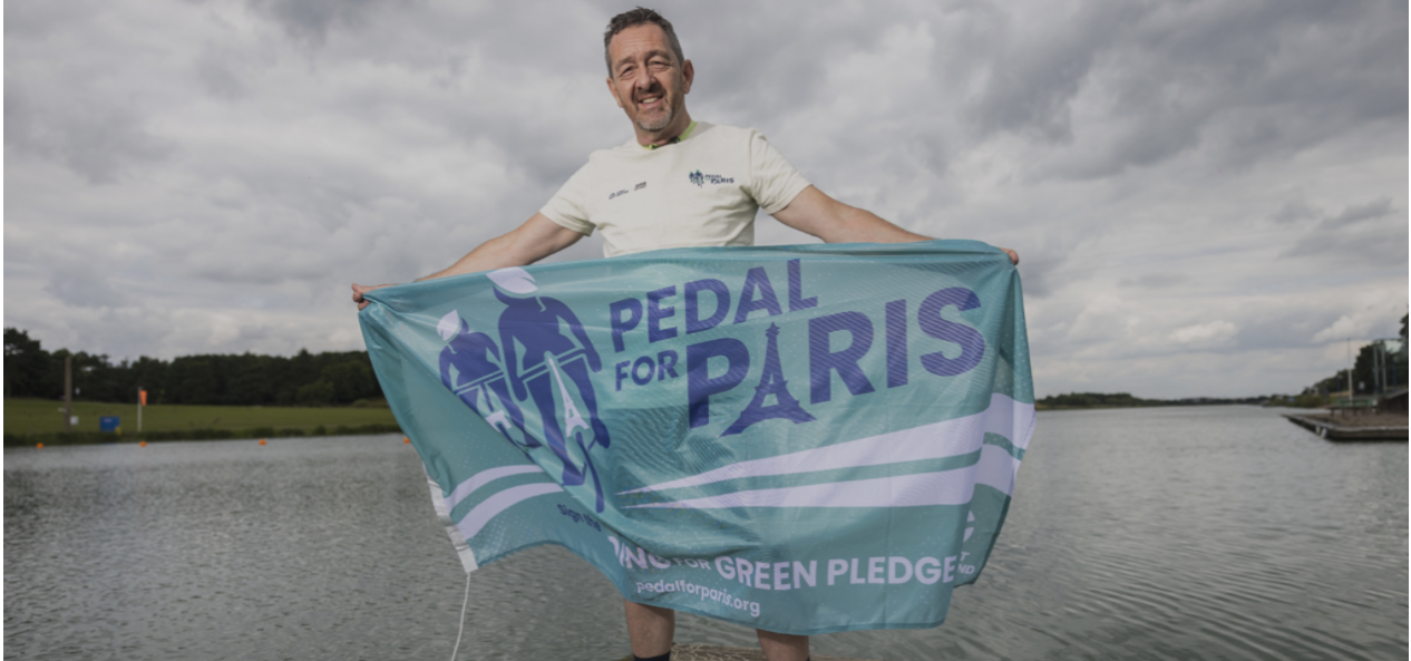 Chris Boardman holds a Pedal for Paris flag in front of a lake